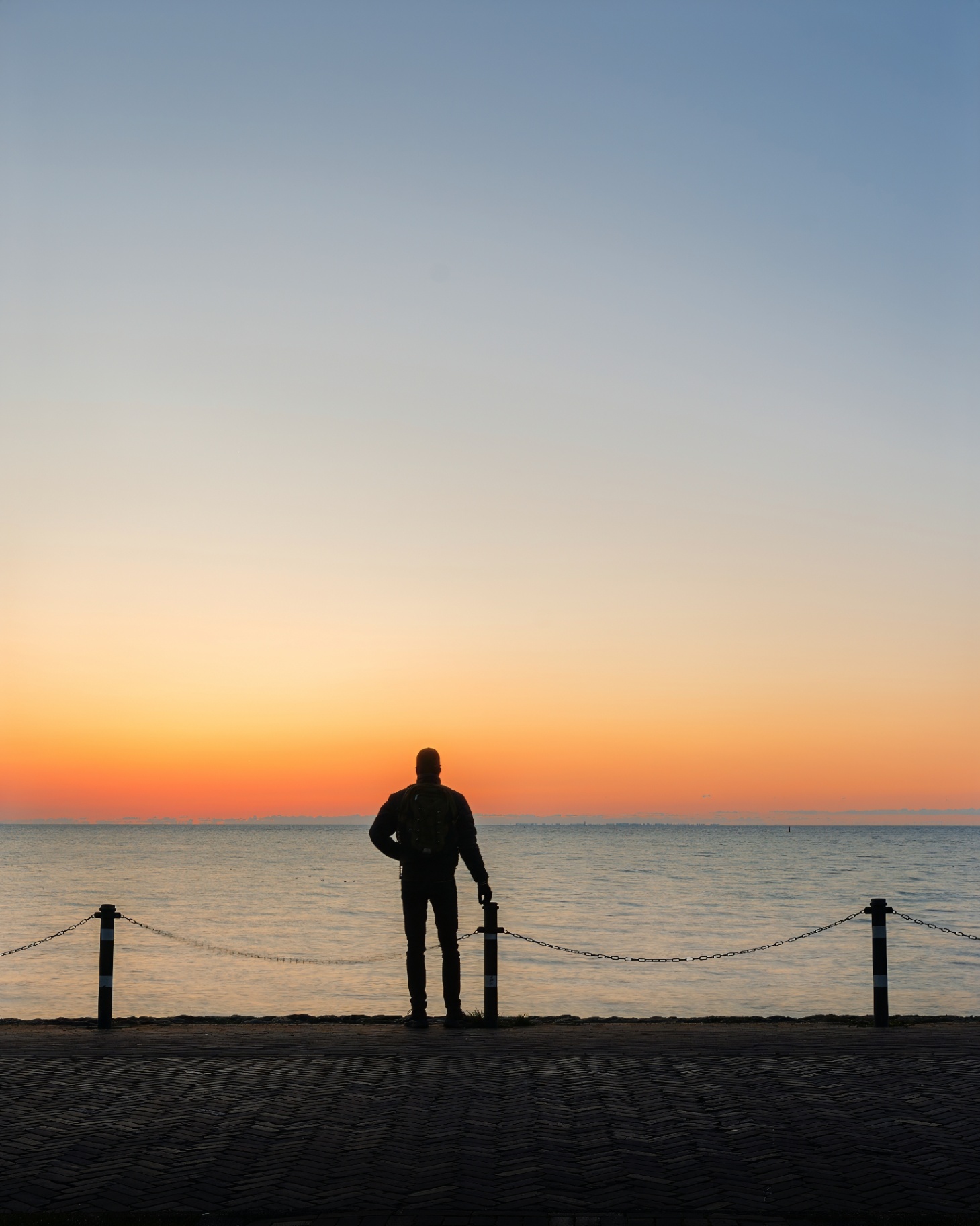 A silhouette watches the sea at first light, standing between two chained posts.