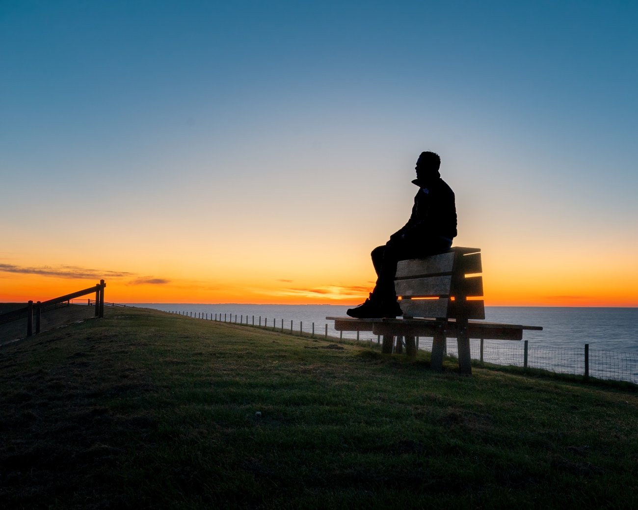 Robin zit op een bankje boven de Waddenzee en kijkt naar een wijde oranje horizon in de schemering.